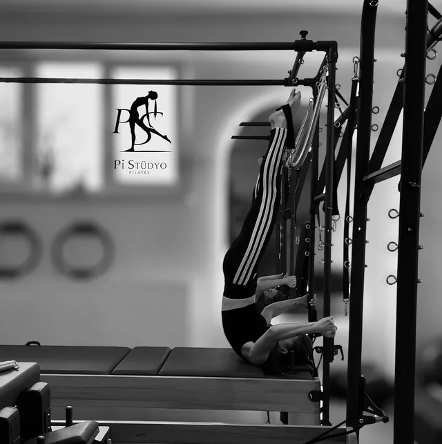 Cadillac üzerinde ters (baş aşağı) pilates egzersizi; pencerede Pi Stüdyo logosu (siyah-beyaz)